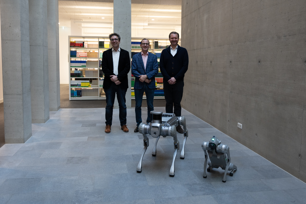 Adrian Merkel, Prof. Dr. Thomas Voit und Prof. Dr. Bartosz von Rymon Lipinski stehen mit den beiden Roboterhunden in der Hochschulbibliothek.